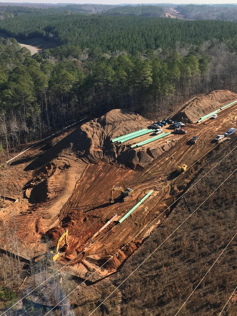 Aerial view of a construction site with heavy machinery and large green pipes placed alongside, surrounded by forested land. Several vehicles are parked near the site.