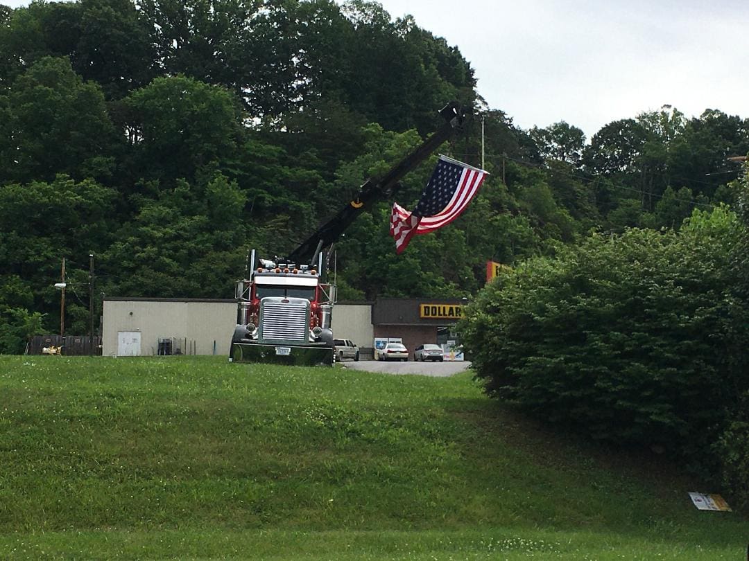 A tow truck with an extended boom displays an American flag outside a Dollar General store, surrounded by greenery and trees.