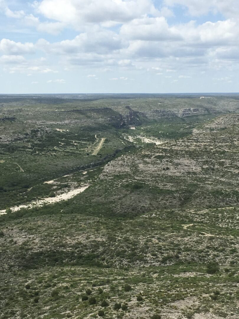 A vast, rocky landscape with sparse vegetation under a partly cloudy sky. A winding dirt road and scattered greenery are visible across the terrain.