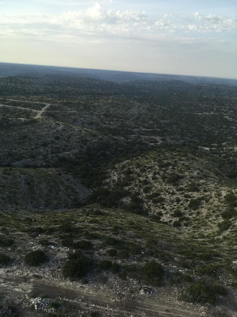 A vast landscape with rolling hills and sparse vegetation under a partly cloudy sky, viewed from a high vantage point. Dirt paths are visible winding through the area.